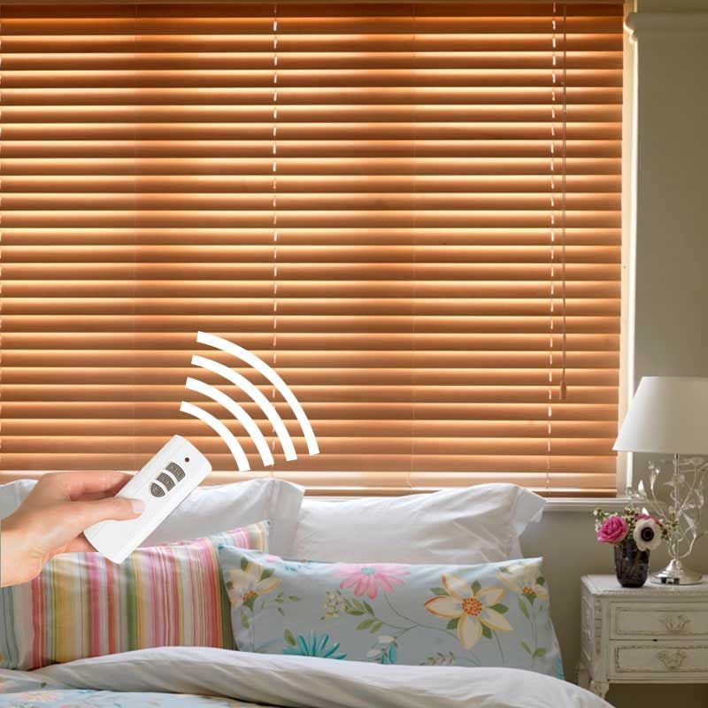A hand aims a white remote at honey-toned wooden venetian blinds over a bed