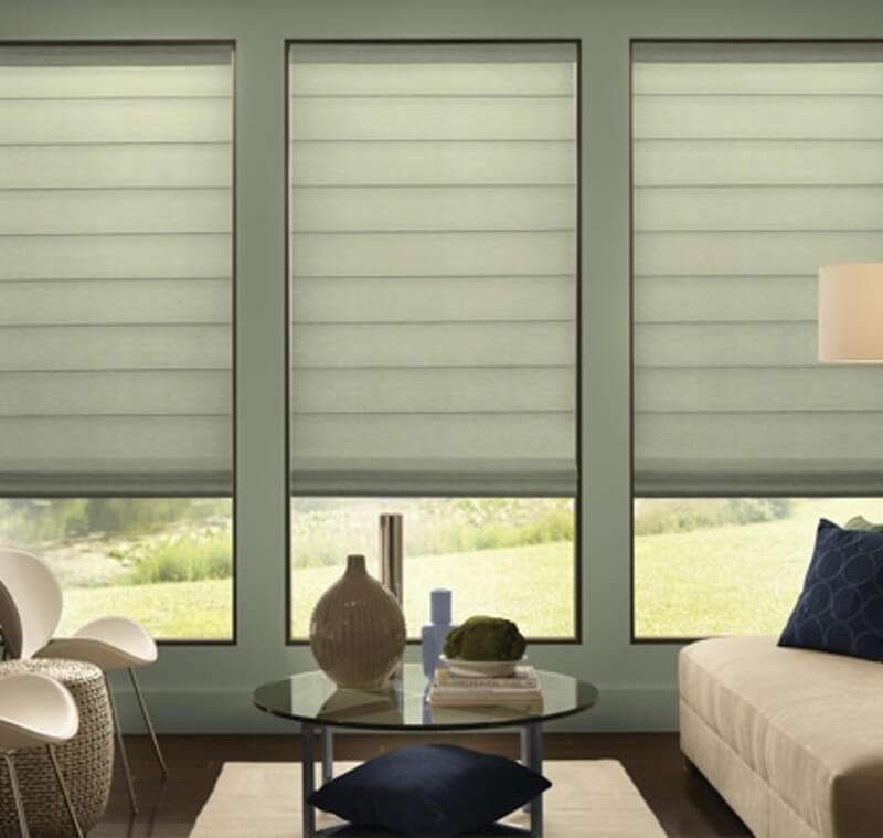 Sage-grey roman blinds lowered three-quarters over three tall windows in a green-walled lounge with a glass table