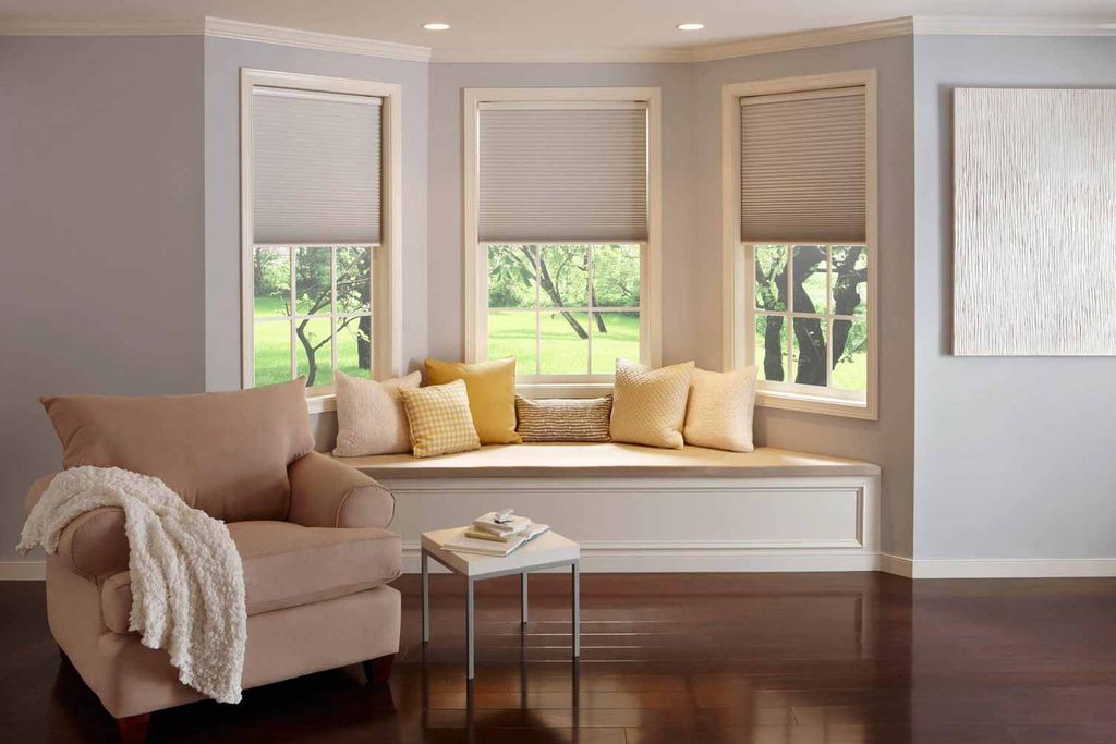 Beige cellular honeycomb blinds half lowered across three bay windows above a cushioned window seat