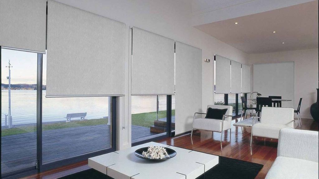 Pale grey roller blinds lowered over floor-to-ceiling glass doors in a waterfront living room, water visible below