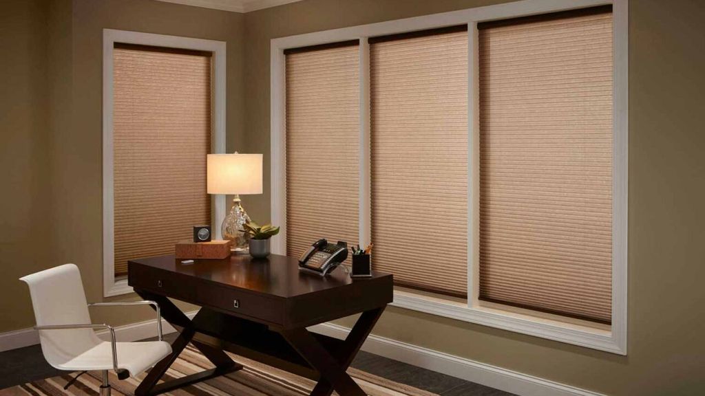 Tan cellular pleated shades closed across four office window panels behind a dark wood desk and white chair