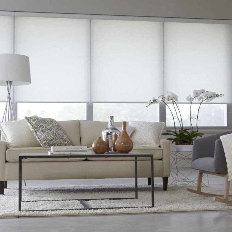 White roller blinds lowered over a wide window wall, leaving a strip of daylight above a cream sofa