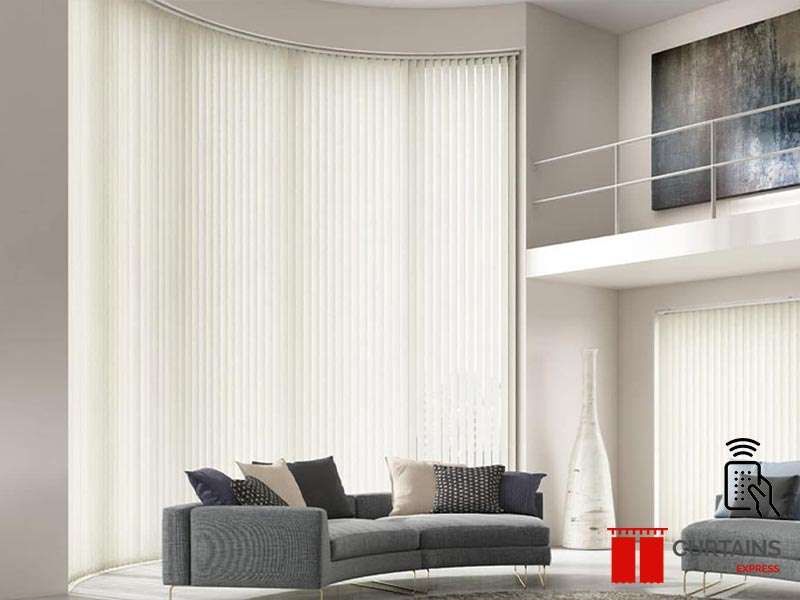 Cream vertical blinds sweeping across a tall curved window wall above a grey curved sofa in a double-height room