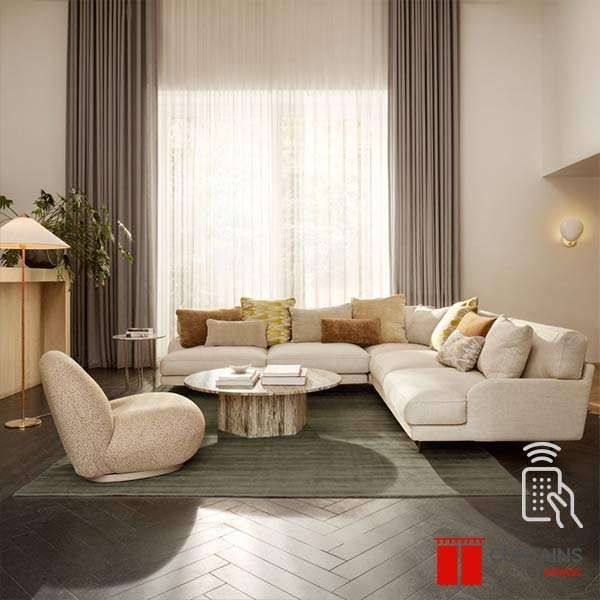 Greige curtains drawn back from a sunlit white sheer behind a cream sectional on a dark herringbone floor