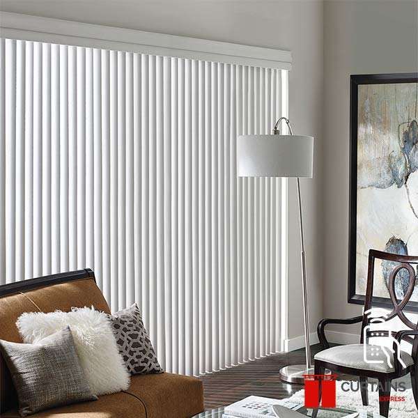 White vertical blinds closed across a patio door, beside a tan sofa and an arc floor lamp