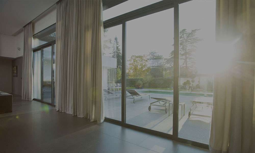 Cream floor-length curtains at sliding glass doors, low sun flaring across a terrace beyond, dark tiled floor