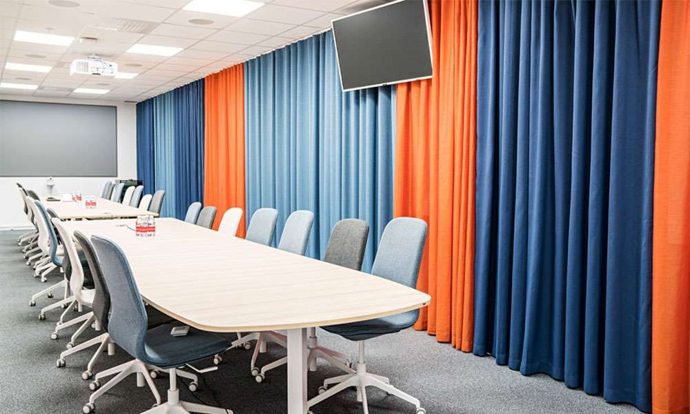 Blue and orange curtains drawn across a meeting room wall, long white table and wheeled chairs alongside