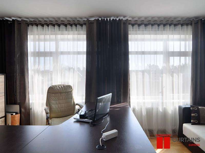 Dark brown eyelet curtains over white sheers on a corner office window, with a laptop on a dark wood desk