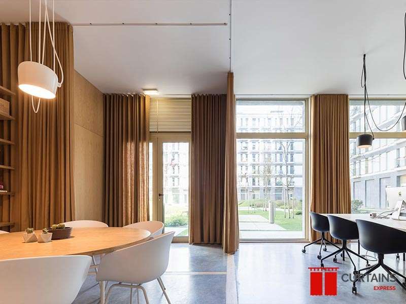 Tan floor-to-ceiling curtains on ceiling tracks along an office window wall, with a wooden slat blind behind