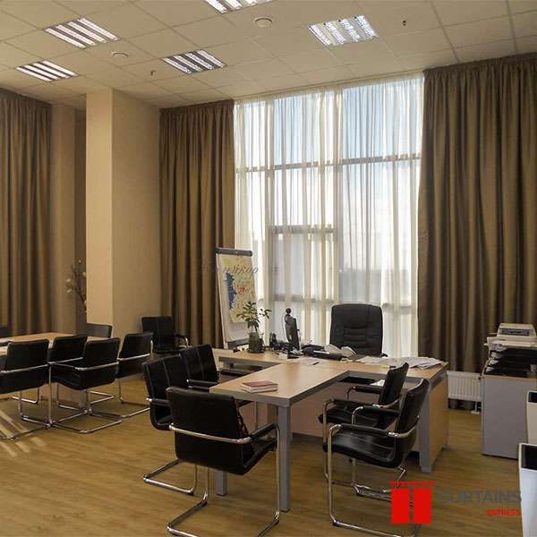 Brown curtains framing a white sheer over corner windows in an office with a desk and black leather chairs