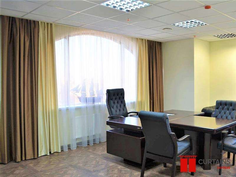 Brown and olive curtains flanking a white sheer at an arch-topped office window above a dark desk and chairs