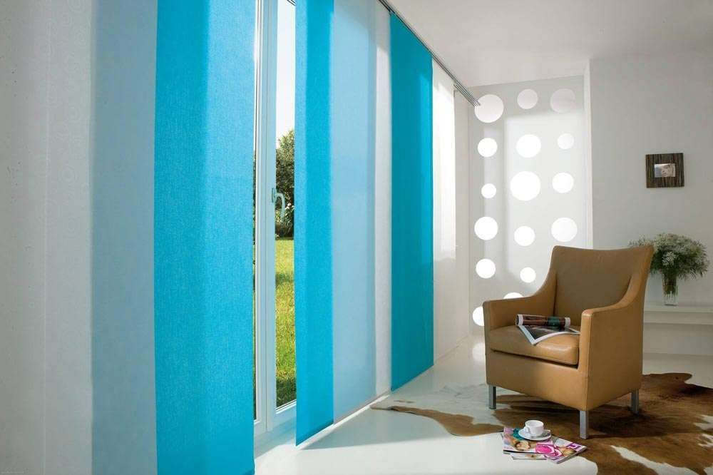 Turquoise sliding panel blinds parted at a balcony door, with a tan leather armchair and cowhide rug beyond