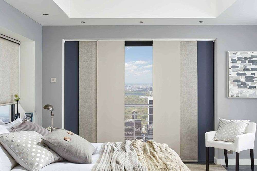 Cream, navy and textured panel blinds across a high-rise bedroom window, left open on a city and park view