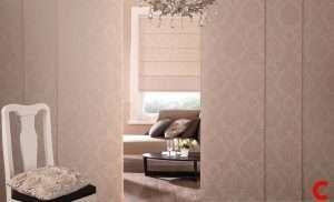 Blush damask sliding panel blinds nearly closed, a gap showing a sofa and cream roman blind behind