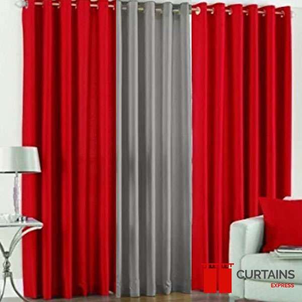 Two red eyelet curtain panels flanking a grey panel on one rod, beside a white armchair