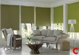 Olive green roller blinds partly lowered across three living room windows behind a grey sofa