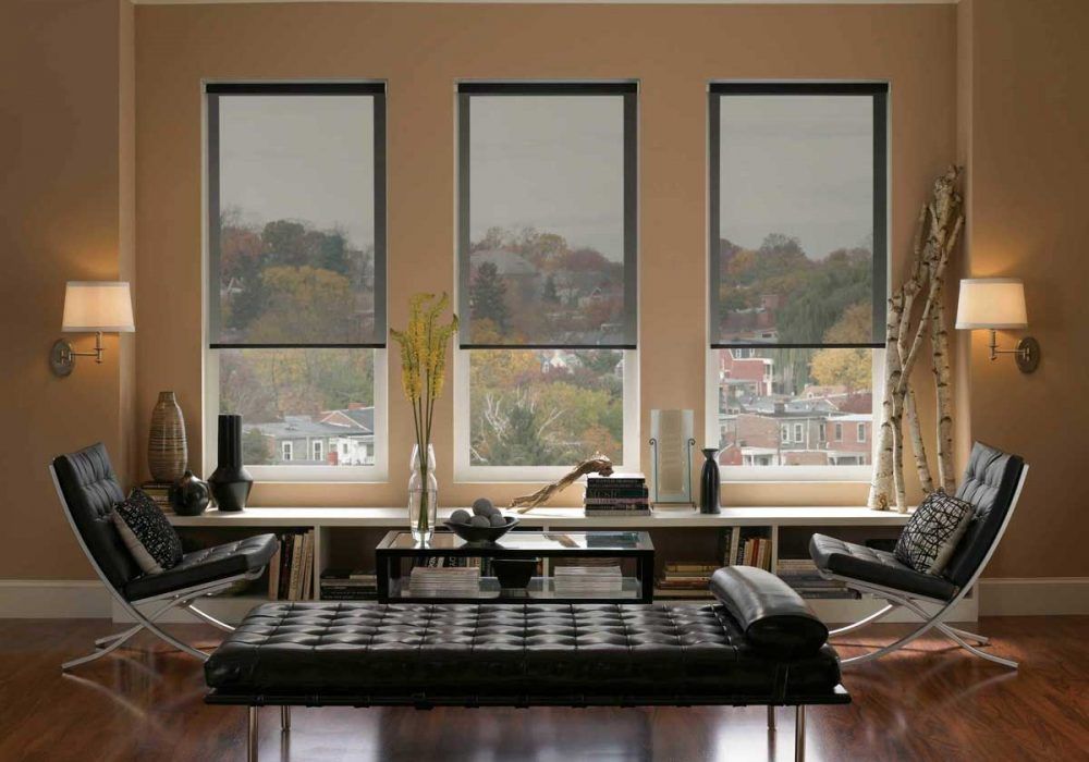 Grey sunscreen roller blinds half lowered over three windows, the treetops still visible through the weave