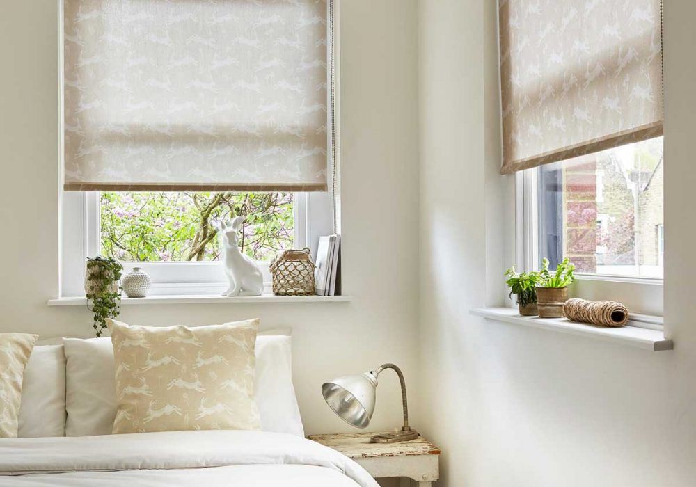 Cream roller blinds printed with white leaping hares, lowered over two bedroom windows above a matching cushion