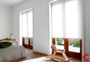 White roller blinds lowered over two tall wood-framed bedroom windows, a rocking horse on the floor