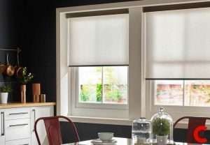 Oatmeal roller blinds over two kitchen windows in a dark-walled room with copper pans and red chairs