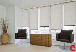 Cream roller blinds two-thirds down a row of five full-height office windows behind brown swivel chairs