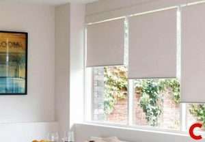 Pale greige roller blinds half lowered over three white-framed windows in a bright white room