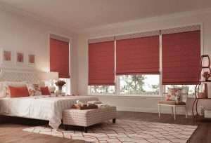 Deep red roman blinds lowered across a run of bedroom windows behind a white bed and padded bench