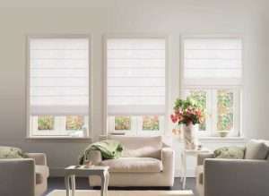 White roman blinds lowered three-quarters over three living room windows above a cream sofa