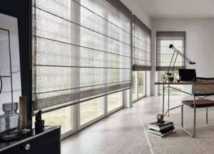 Grey open-weave roman blinds along a floor-to-ceiling window wall in a home office with a desk lamp