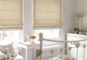 Cream striped roman blinds at two corner windows in a nursery with a white iron bed and radiator
