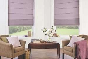 Heather purple roman blinds across a three-sided bay window between two beige armchairs
