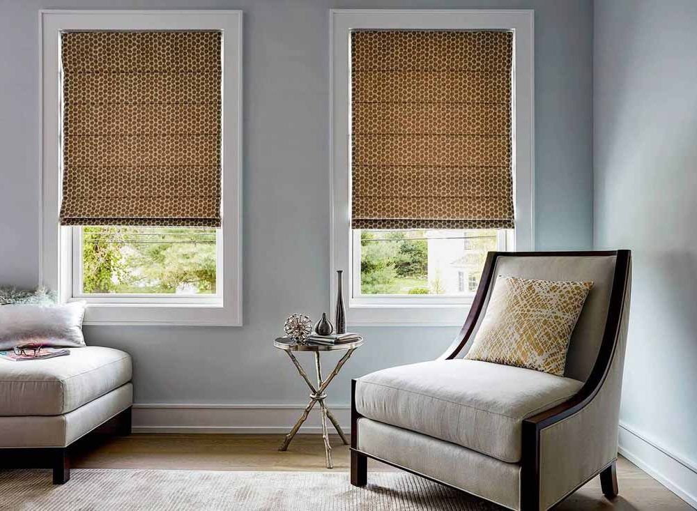 Tan circle-patterned roman blinds lowered over two windows in a pale blue room with a cream armchair