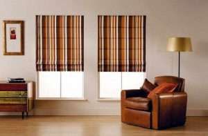 Brown and orange striped roman blinds over two windows beside a tan leather armchair and floor lamp
