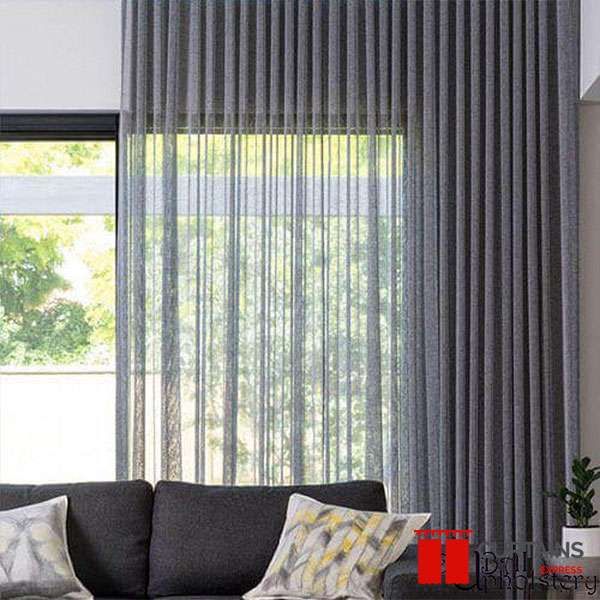 Charcoal sheer curtains hung ceiling to floor across a corner window, garden trees showing through the loose weave