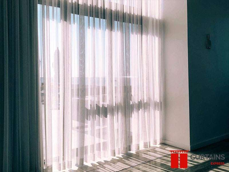 White sheer curtains across a floor-to-ceiling balcony door in strong sunlight, a teal wall and darker panel alongside