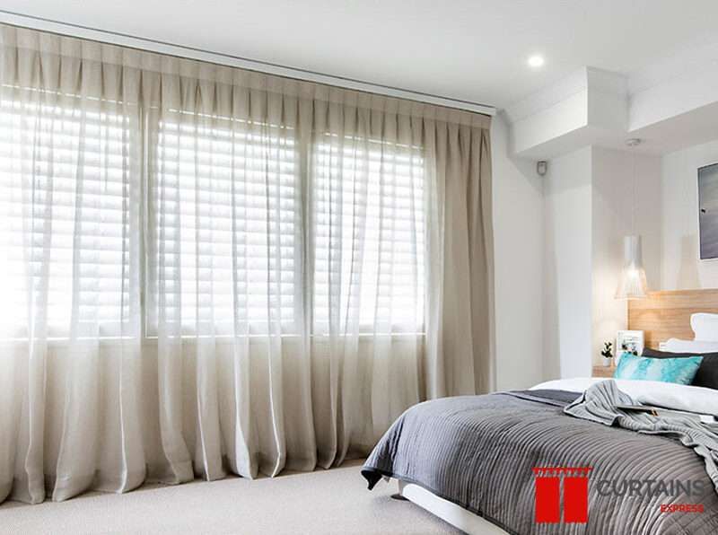 Taupe sheer curtains on a ceiling track spanning a bedroom window, white plantation shutters closed behind them