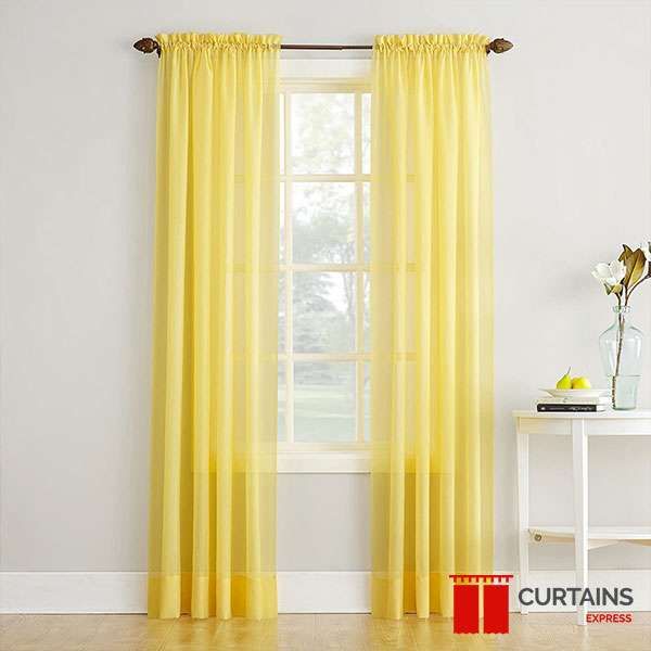 Yellow sheer rod-pocket panels on a bronze pole either side of a small white sash window in a grey-walled room