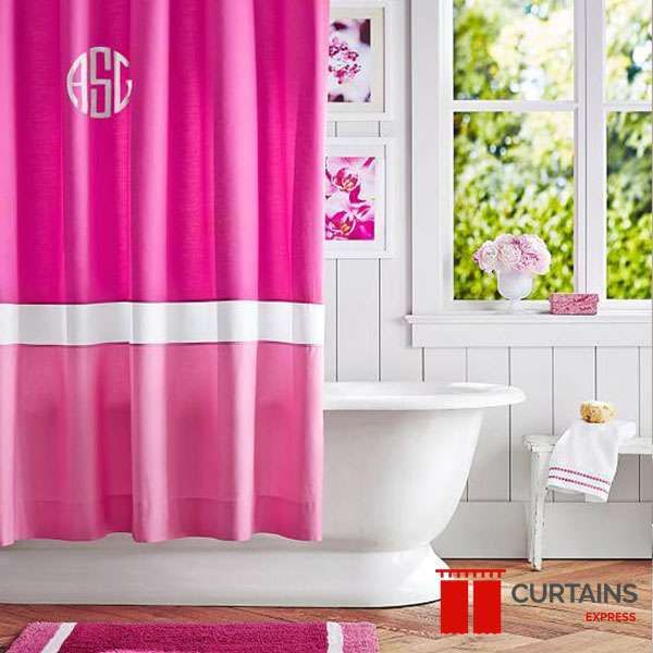 Two-tone pink monogrammed shower curtain with a white band, beside a white roll-top bath and panelled wall
