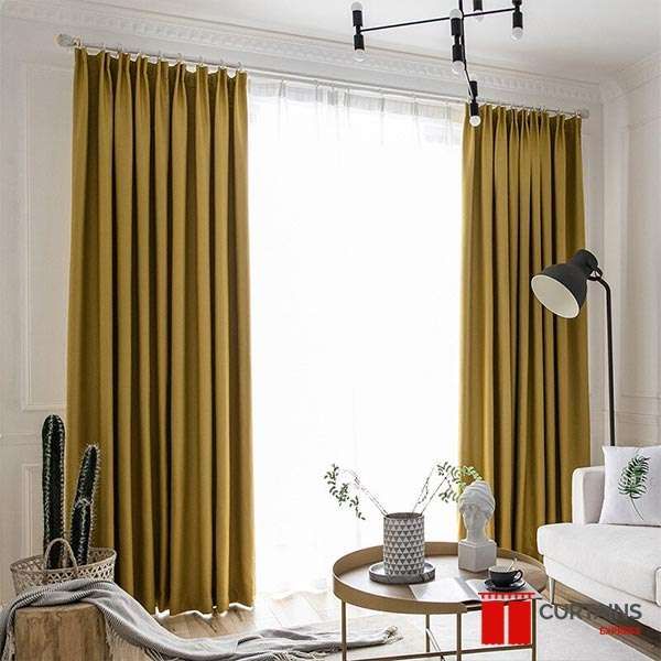 Mustard pinch-pleat curtains on a white pole across a full-height living room window, sheer drawn behind