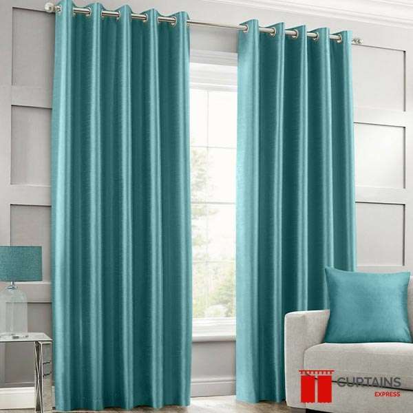 Teal eyelet curtains on a chrome pole at a bay window in a grey panelled room, matching cushion on the armchair