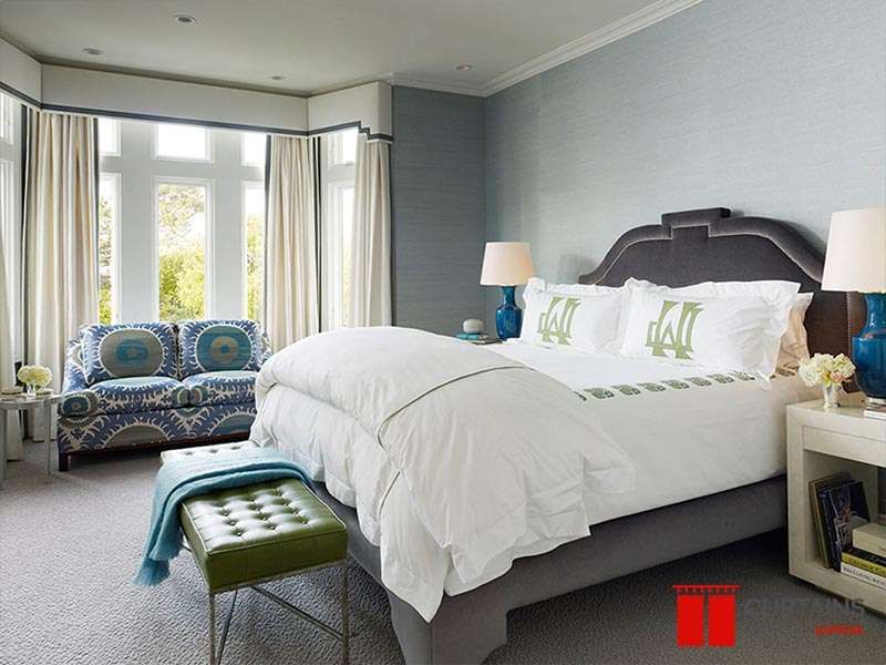 Cream curtains under a navy-trimmed pelmet around a bedroom bay window, patterned blue settee set in the bay