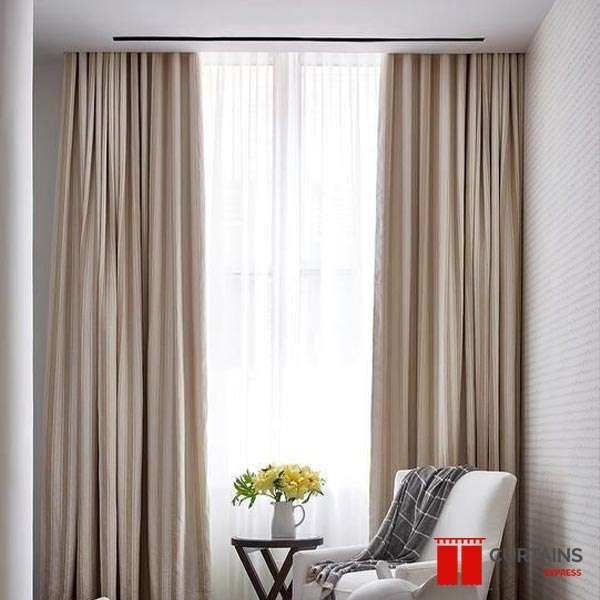 Beige pinch-pleat curtains on a recessed ceiling track layered over white sheers beside a white armchair