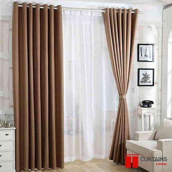 Tan eyelet curtains over white sheers, the right panel gathered by a fabric tieback beside a cream sofa