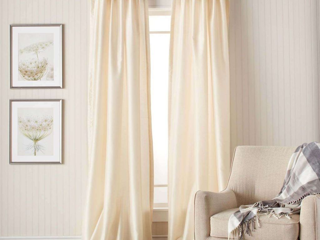 Ivory silk curtains hanging full-length either side of a small window, beside framed botanical prints and a beige armchair