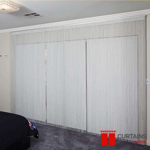 White string curtains hung wall-to-wall across a bedroom window run, with a dark bed and grey carpet in the foreground