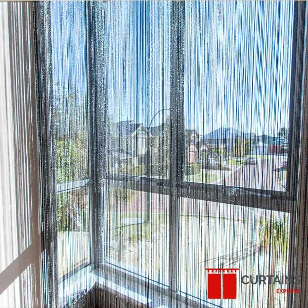 Charcoal string curtains hanging inside a corner window, sunlit suburban rooftops and hills visible through the strands
