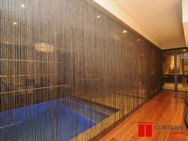 Bronze string curtains screening an indoor pool from a timber-floored hallway, lit blue water behind the strands
