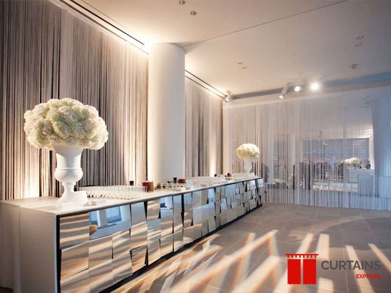 Beige string curtains lining the walls of an event room behind a long mirrored bar counter topped with white flowers