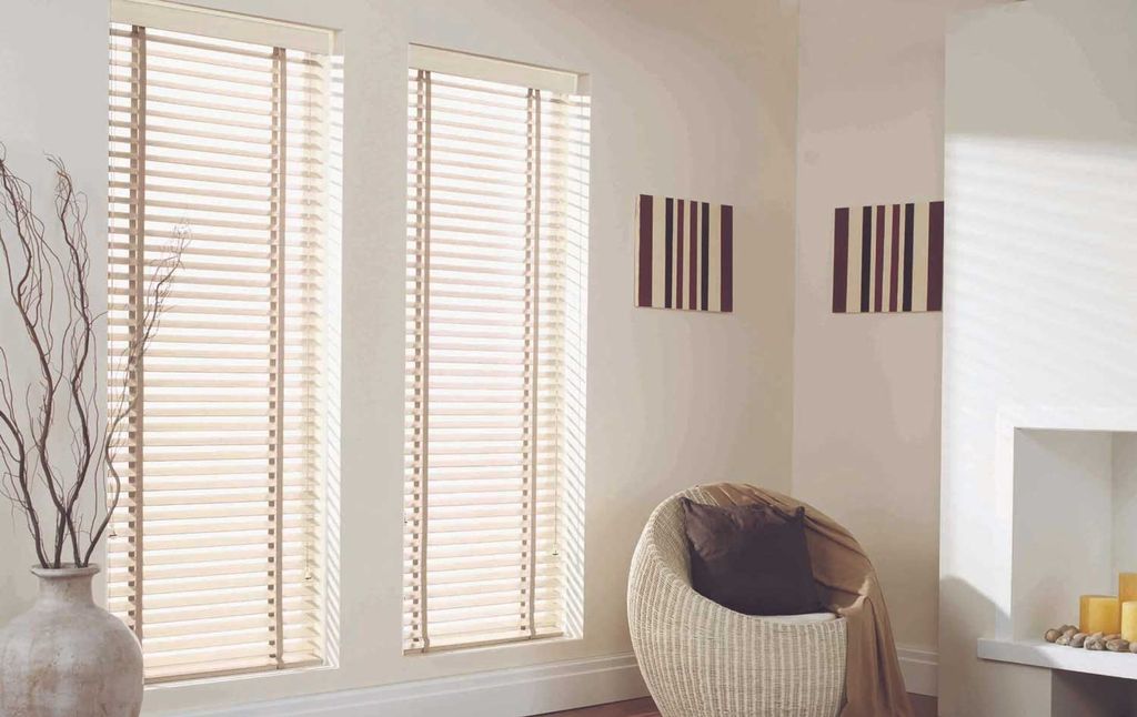 Cream wide-slat venetian blinds tilted almost closed on two tall windows, sunlight edging every slat