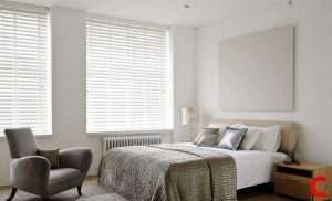 White venetian blinds across a wide bedroom window, with a grey armchair, radiator and knitted bed throw below